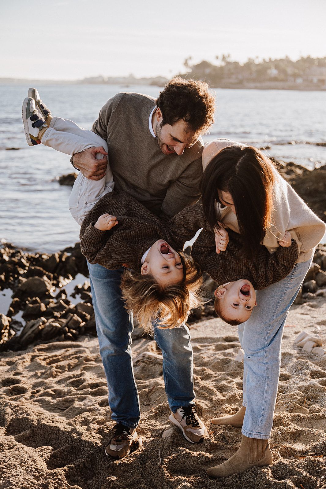 Family playfully holding children upside down on a sandy beach at sunset.