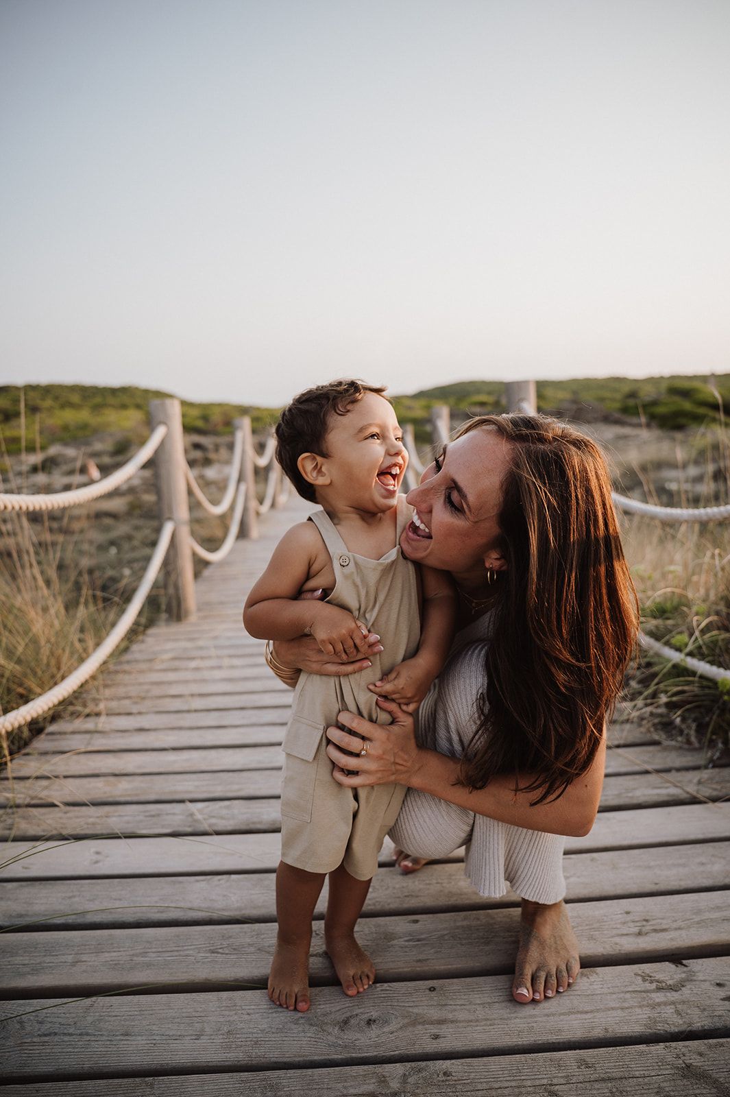 Mother crouching on a wooden boardwalk hugging her laughing toddler at the beach.