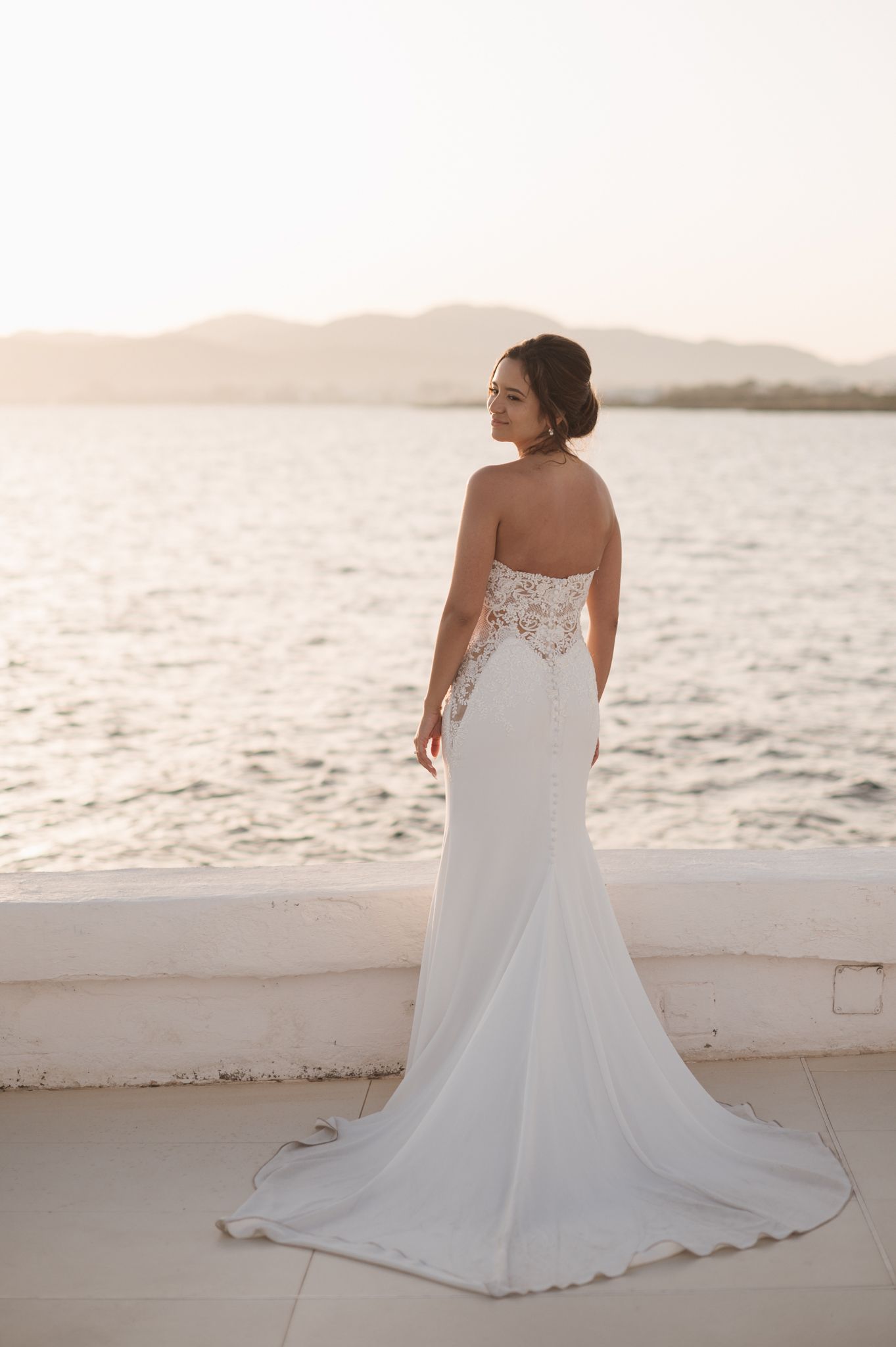 Elegant bride in strapless lace back wedding gown poses by the sea at golden sunset.