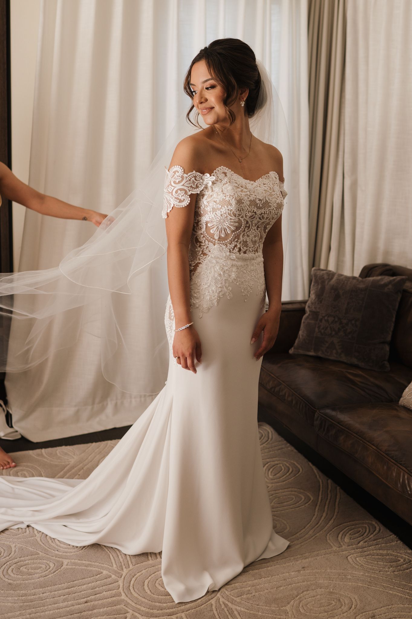 Bride in off-shoulder lace mermaid wedding gown with veil being adjusted in elegant hotel room.