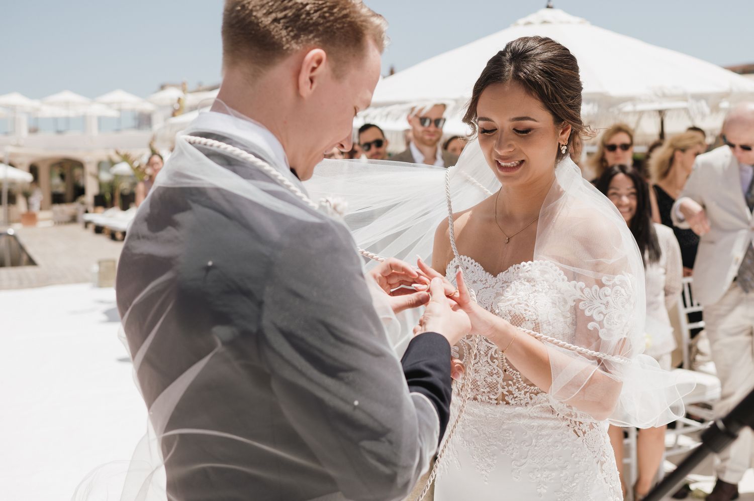 Groom places wedding ring on brides finger during outdoor ceremony in bright sunlight with guests watching.