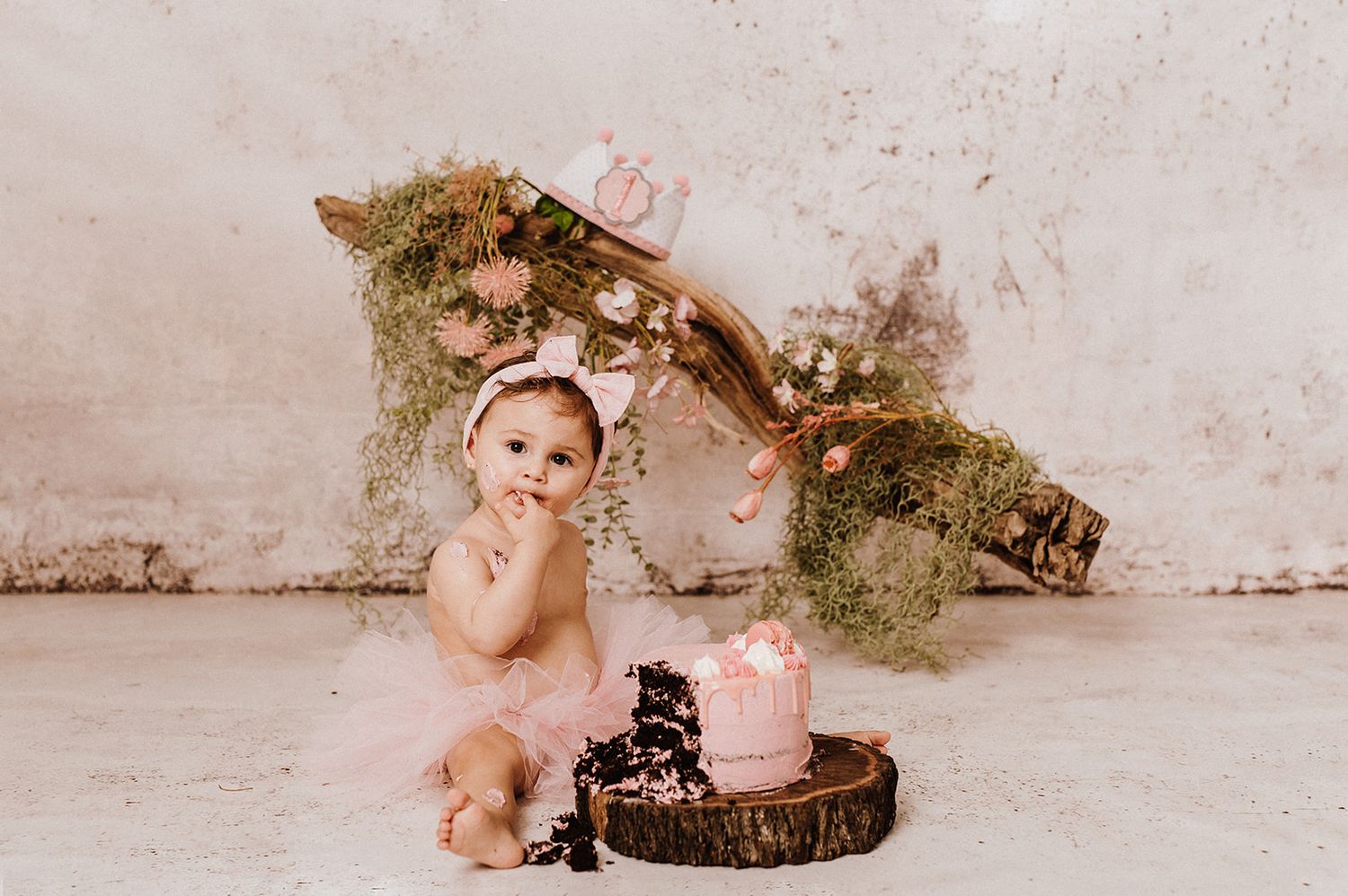 Baby girl in pink tutu sits beside a pink smash cake on a wood slice for her first birthday photo.