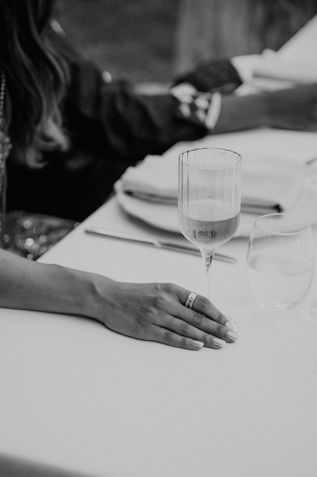 Black and white close-up of a wedding guest's hand with rings resting on a white reception table.