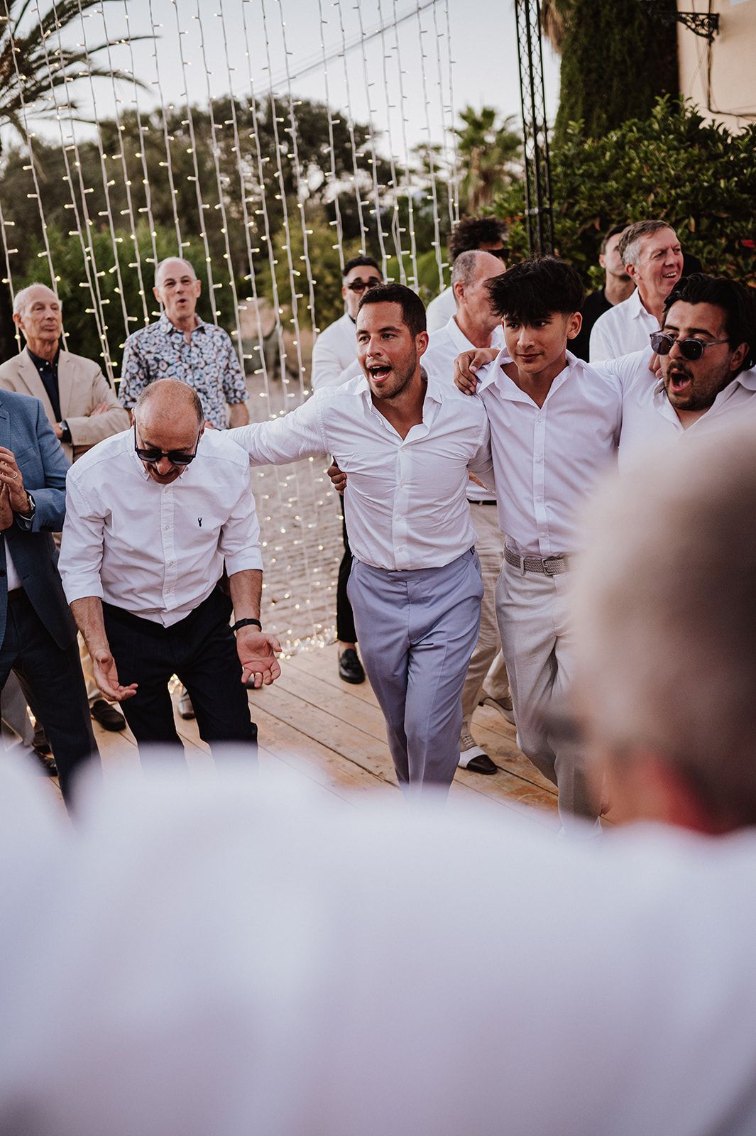 Guests in white attire celebrate joyfully outdoors at a wedding reception with string lights backdrop.