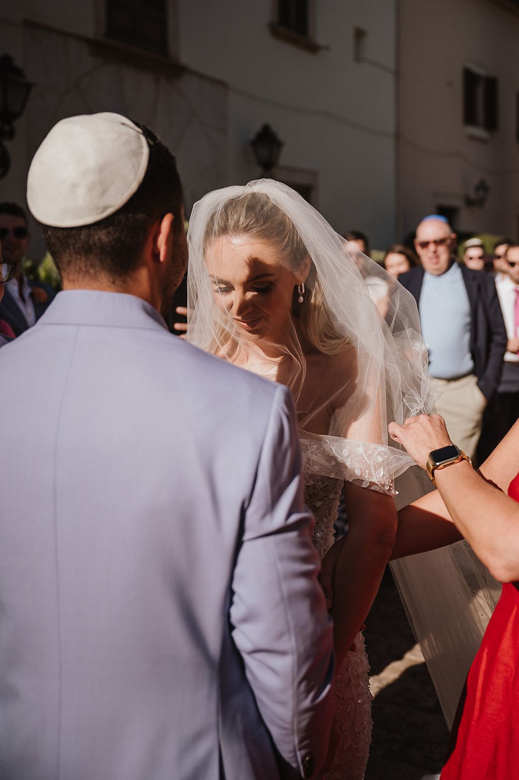 Bride in lace gown and veil during Jewish wedding ceremony with groom in lavender suit wearing kippah.