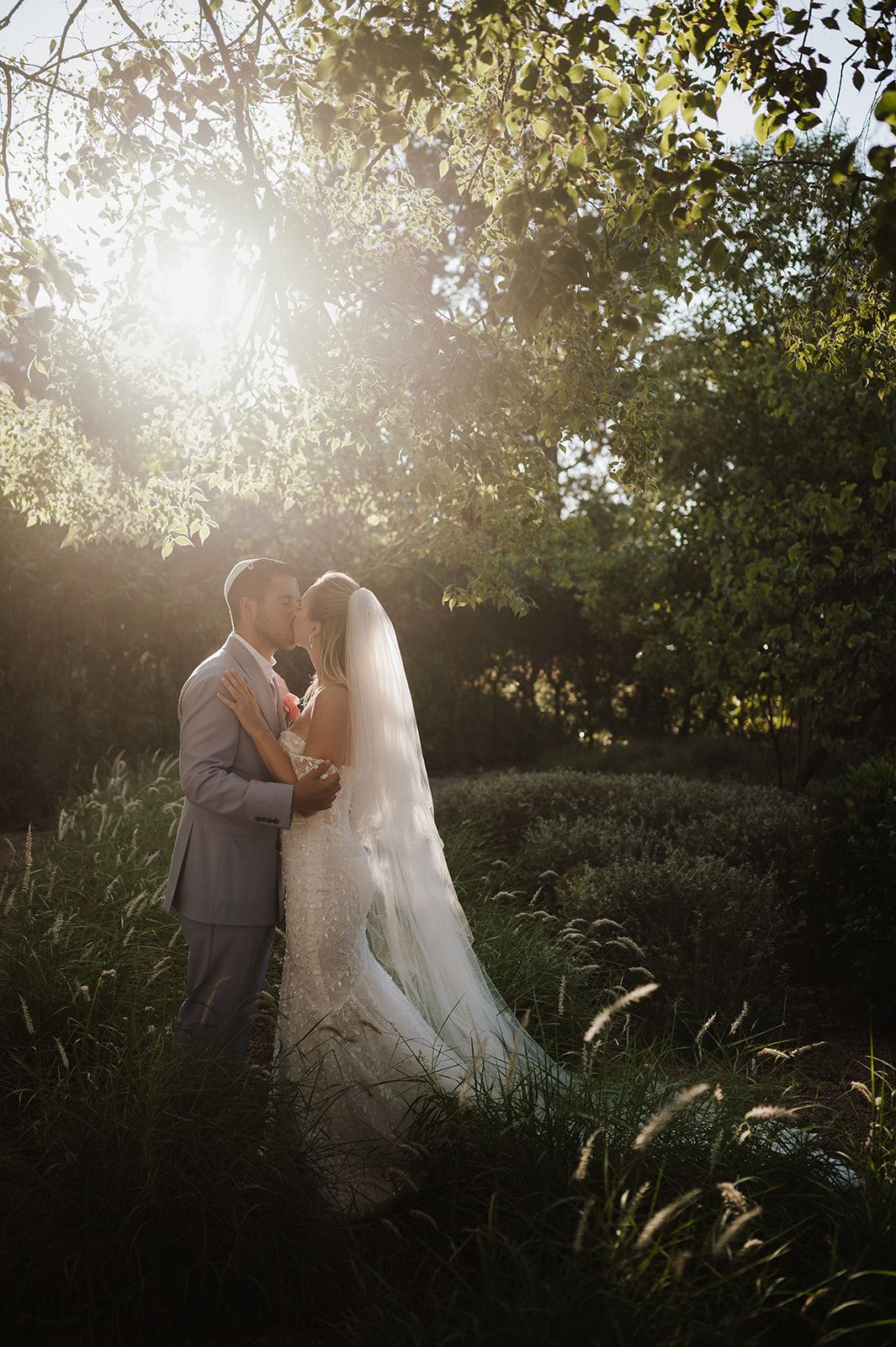 Bride and groom share a romantic kiss surrounded by lush greenery with golden sunlight streaming through the trees.