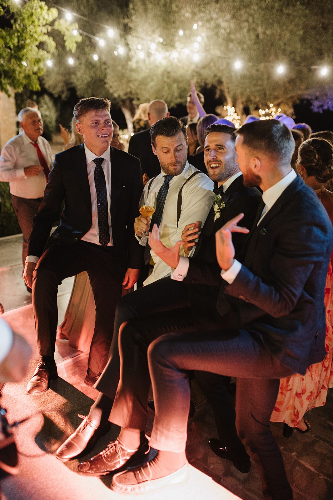 Group of groomsmen laughing and celebrating together under string lights at an outdoor evening wedding reception.