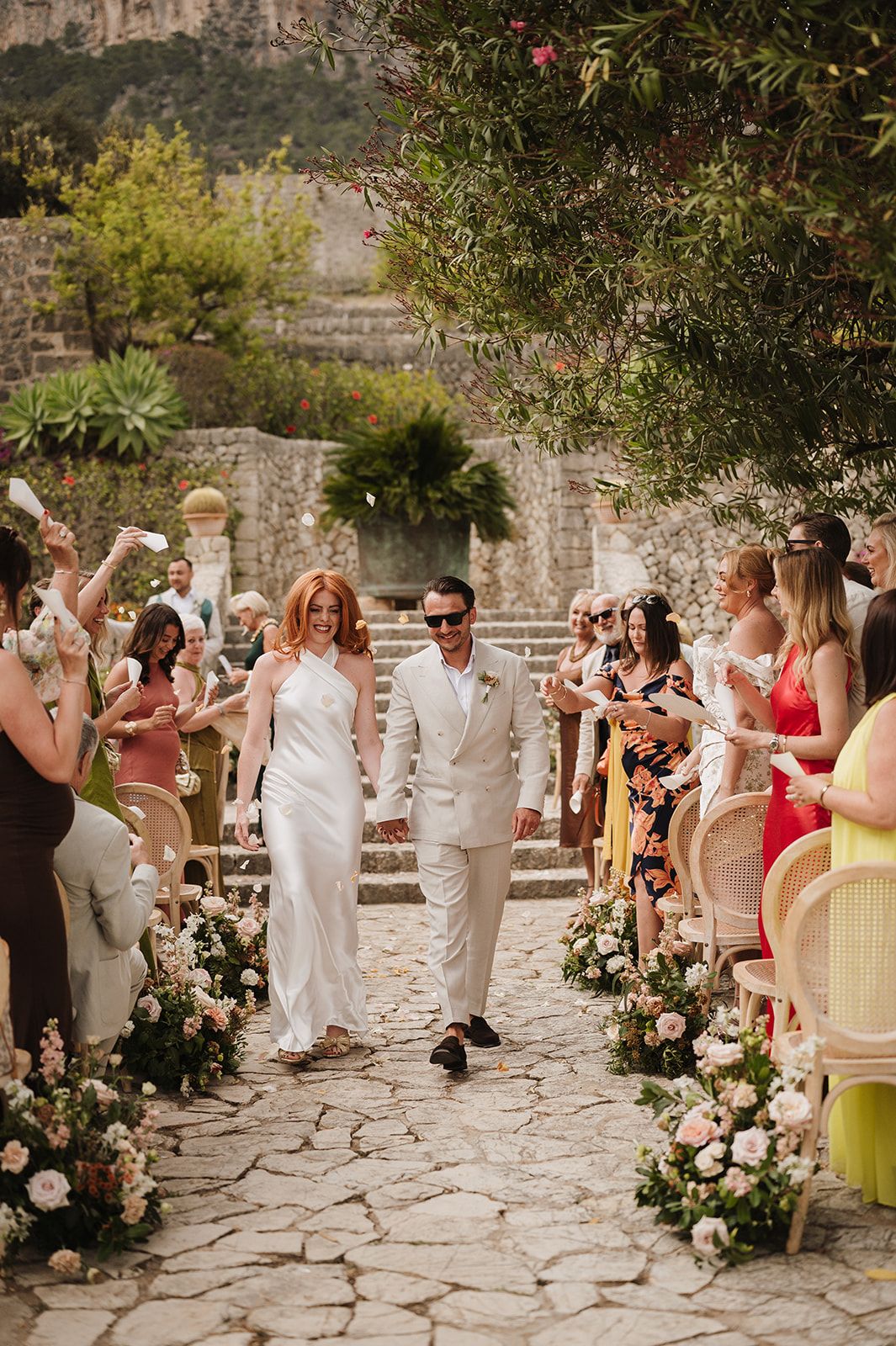 Joyful newlyweds walk through confetti-throwing guests down flower-lined cobblestone aisle after outdoor wedding ceremony.
