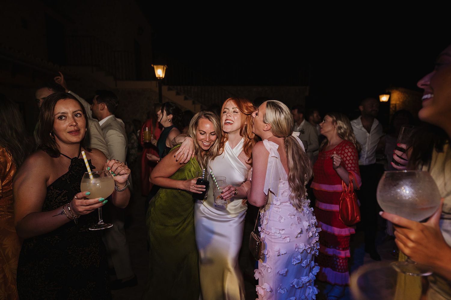 Women in elegant dresses laughing and celebrating together at an outdoor wedding reception.