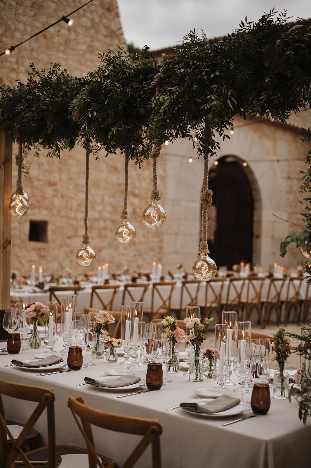 Rustic outdoor wedding reception tables adorned with hanging globe lights, greenery canopy, and pink floral centerpieces.