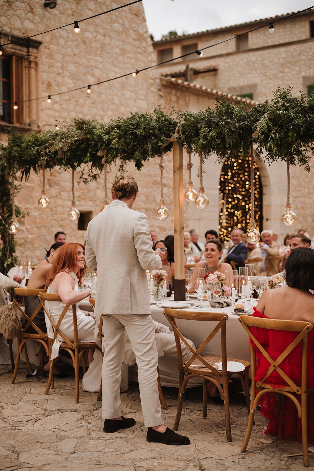 Guest giving a wedding speech at an outdoor stone venue decorated with greenery and Edison bulb lights.
