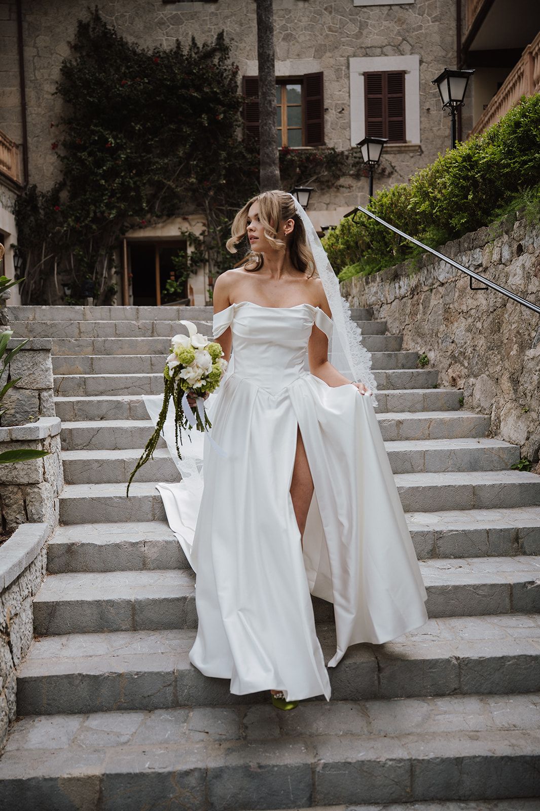 Bride in flowing off-shoulder white dress with veil carrying bouquet on stone staircase.