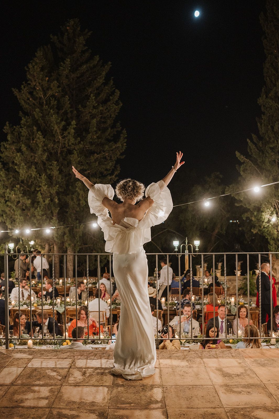 Groom lifts bride in white dress during outdoor wedding reception under string lights and moonlit sky.
