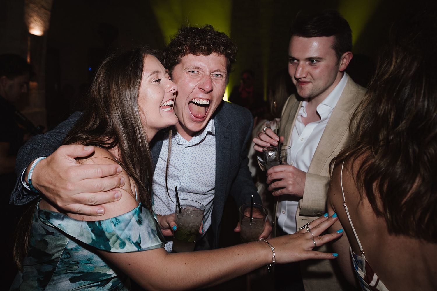Joyful wedding guests laugh and embrace on the dance floor during a lively indoor wedding reception.