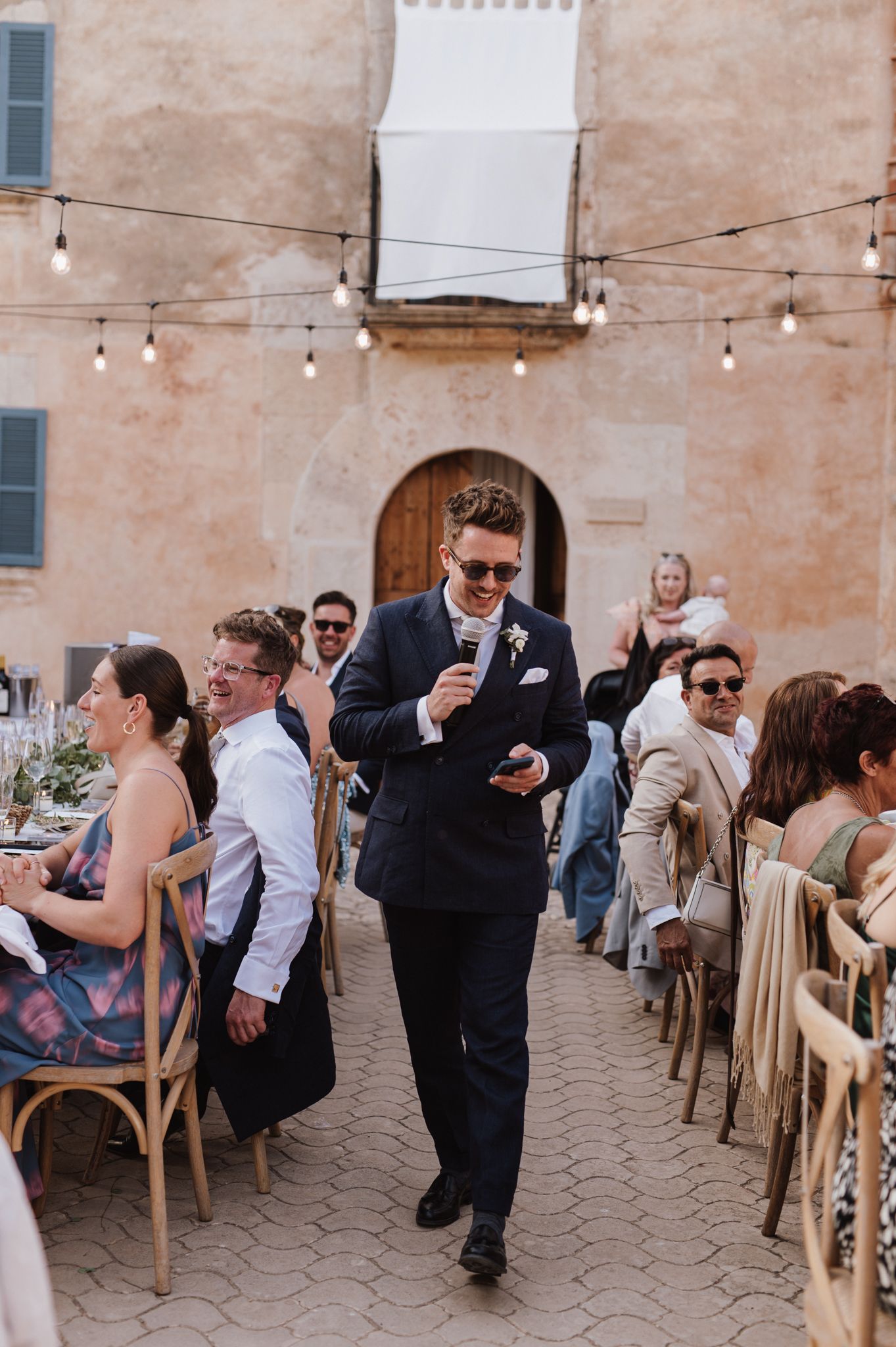 Groom in navy suit giving a speech with microphone at an outdoor wedding reception.