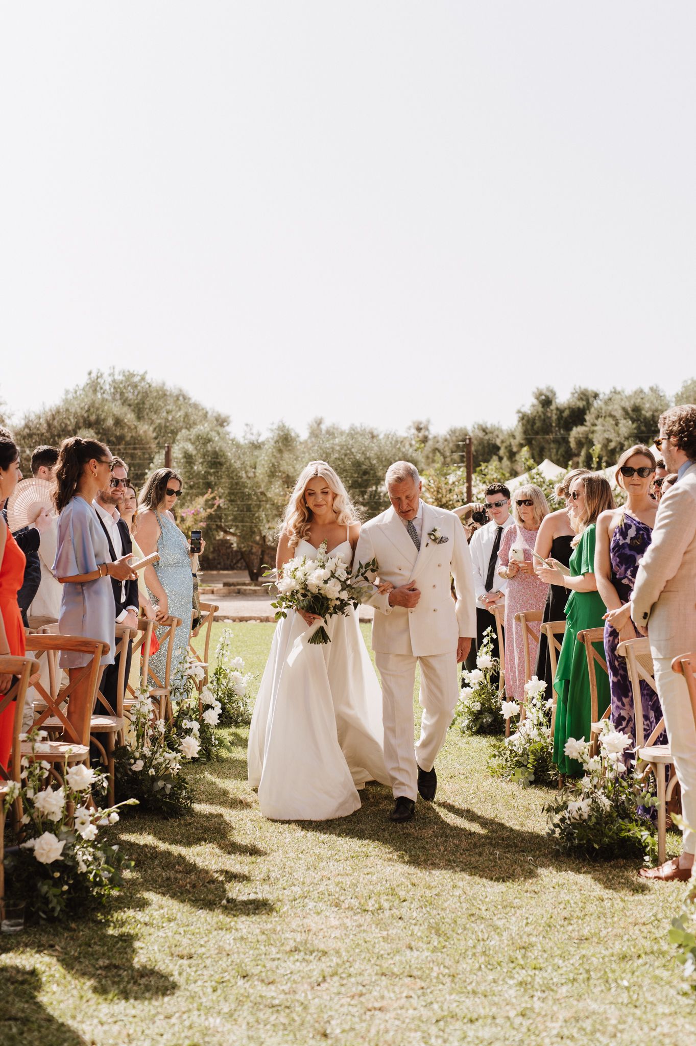 Bride walks down outdoor wedding aisle with escort, flanked by guests and floral arrangements on sunny day.