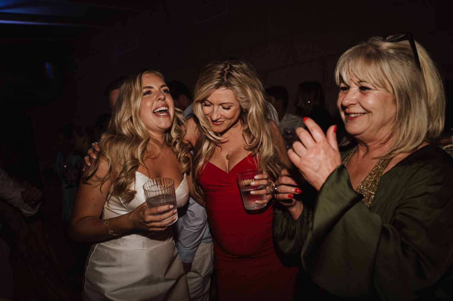 Bride in white gown laughing with two female guests at lively indoor wedding reception party.