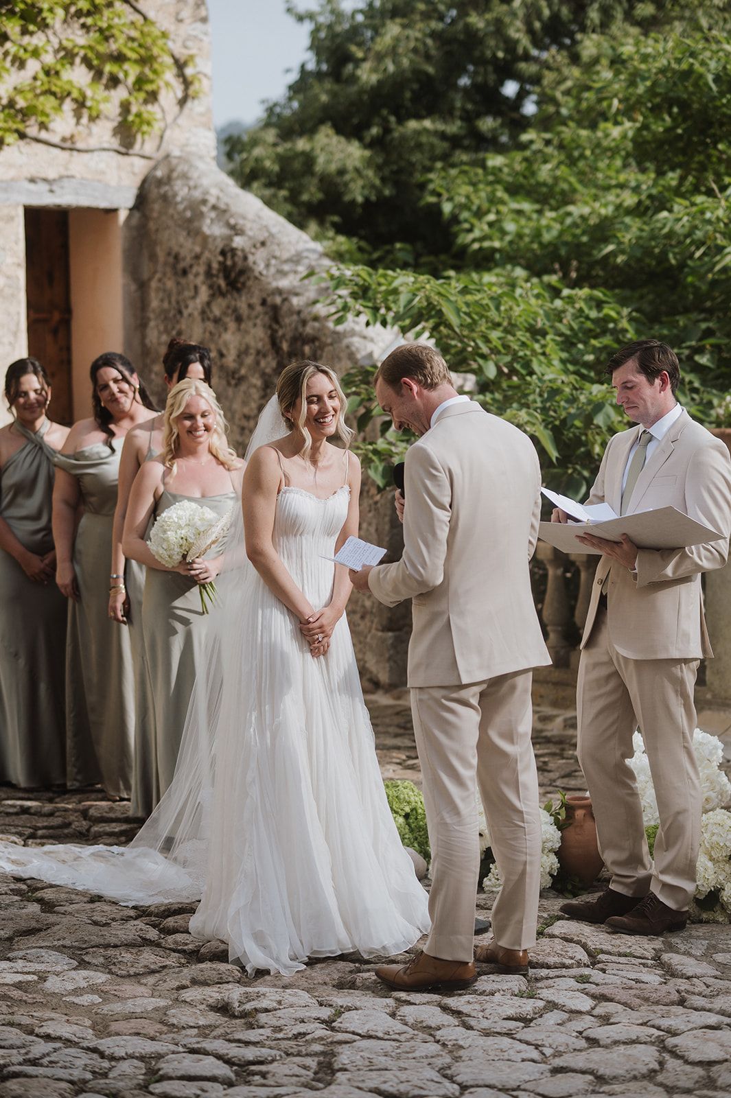Outdoor wedding ceremony on cobblestone with bride in white gown, groom in tan suit reading vows, bridesmaids in sage dresses.