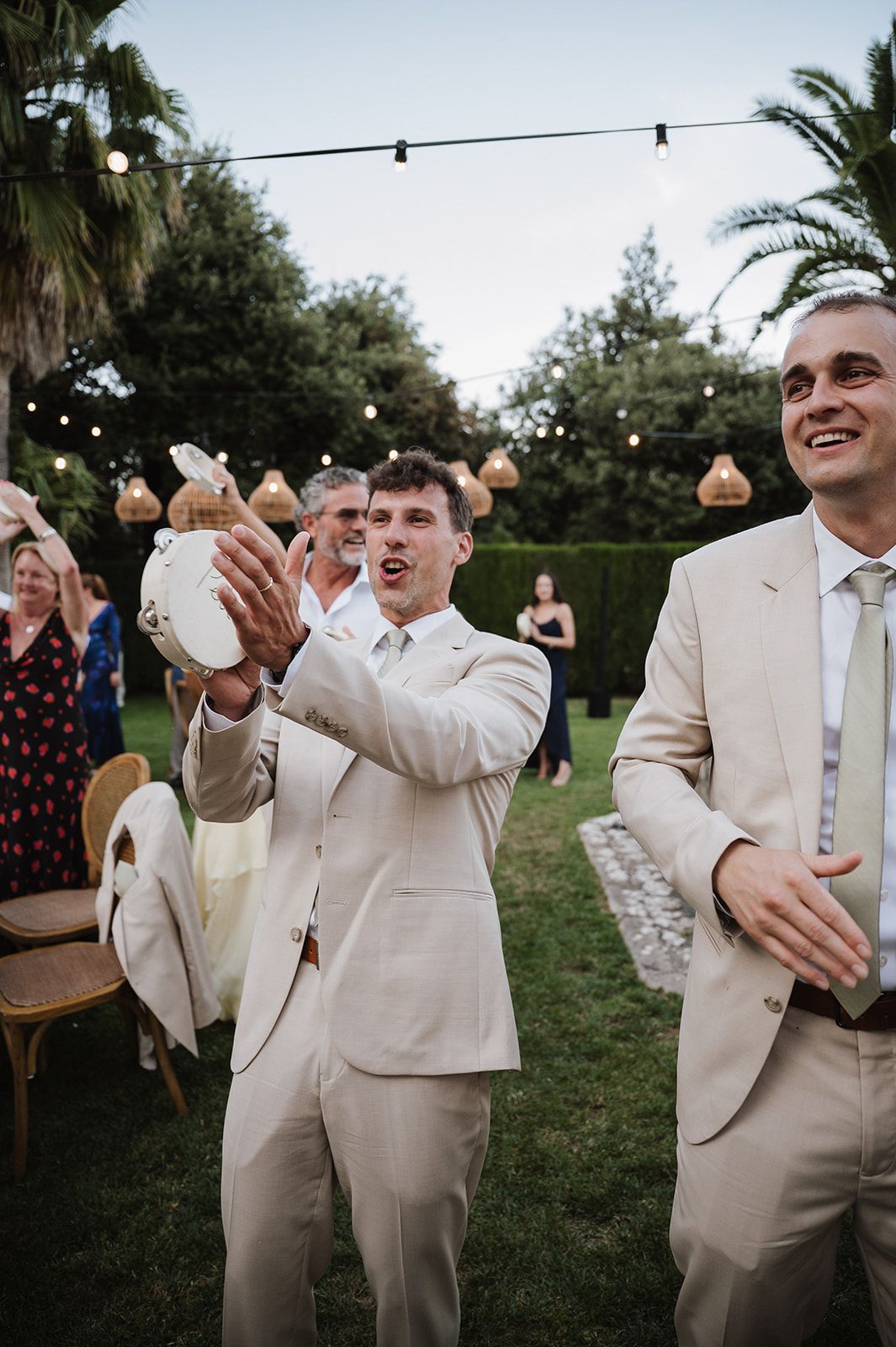 Groomsman in beige suit catches white object at outdoor wedding reception with string lights overhead.