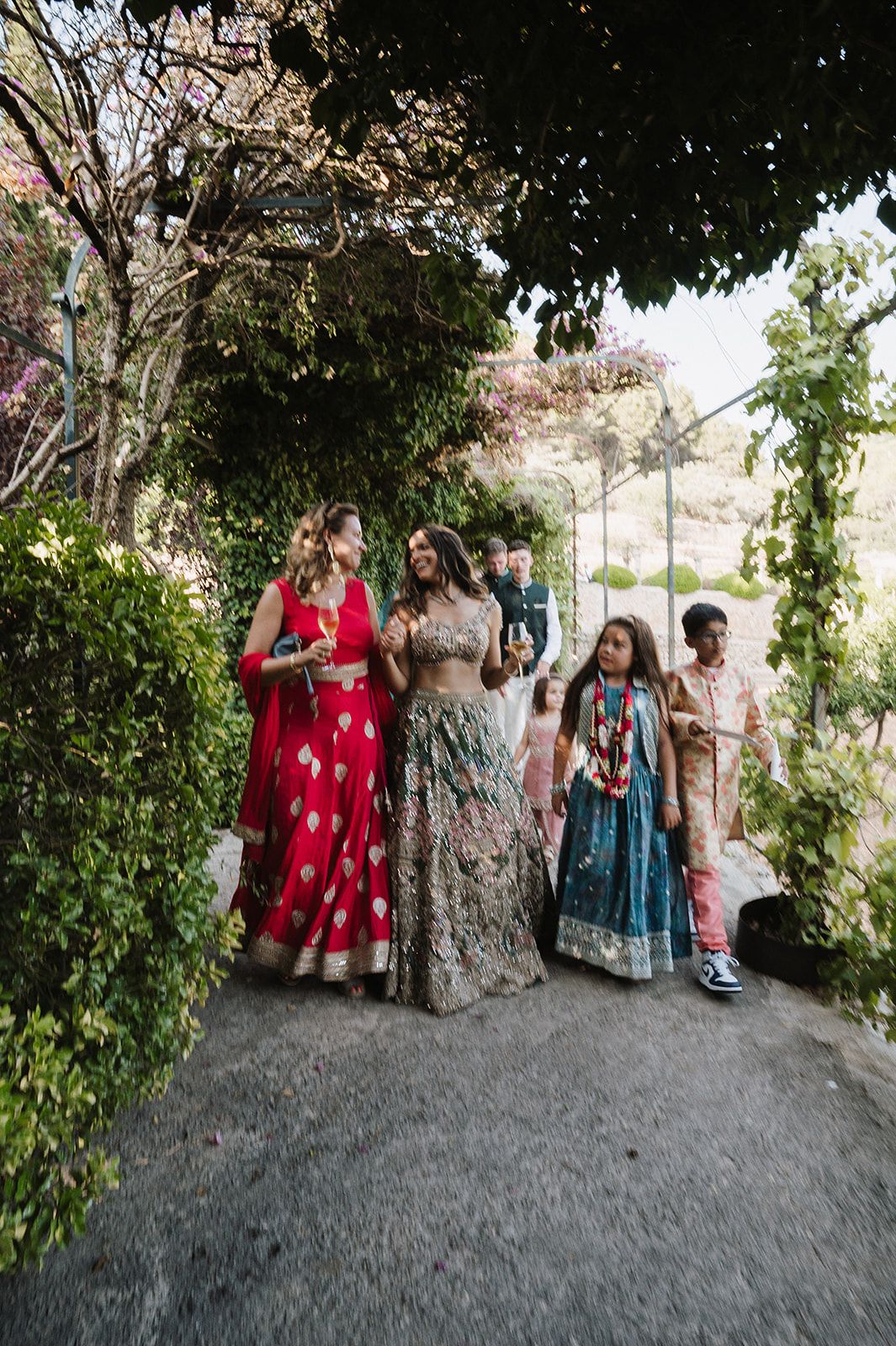 Bride in floral lehenga walks with guests in red and teal Indian attire through a lush garden path.