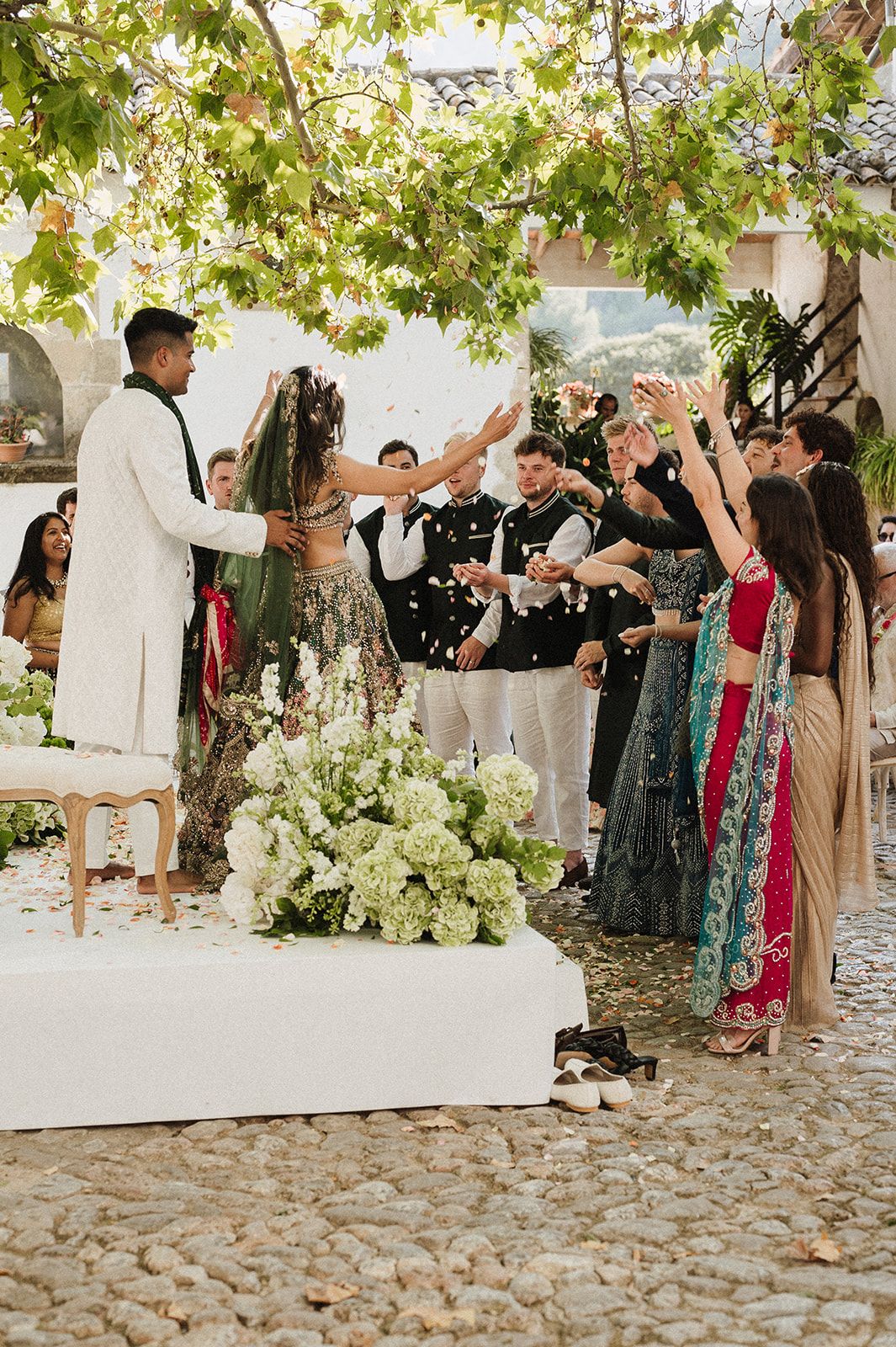 Couple at Hindu wedding ceremony with guests throwing flower petals under canopy of green trees.
