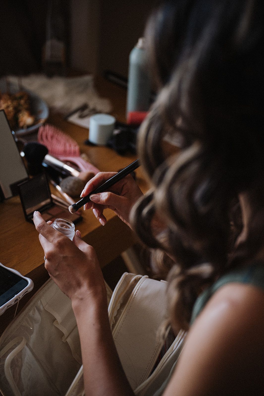 Woman with curly hair applying makeup at a vanity table with beauty tools and products in soft lighting.