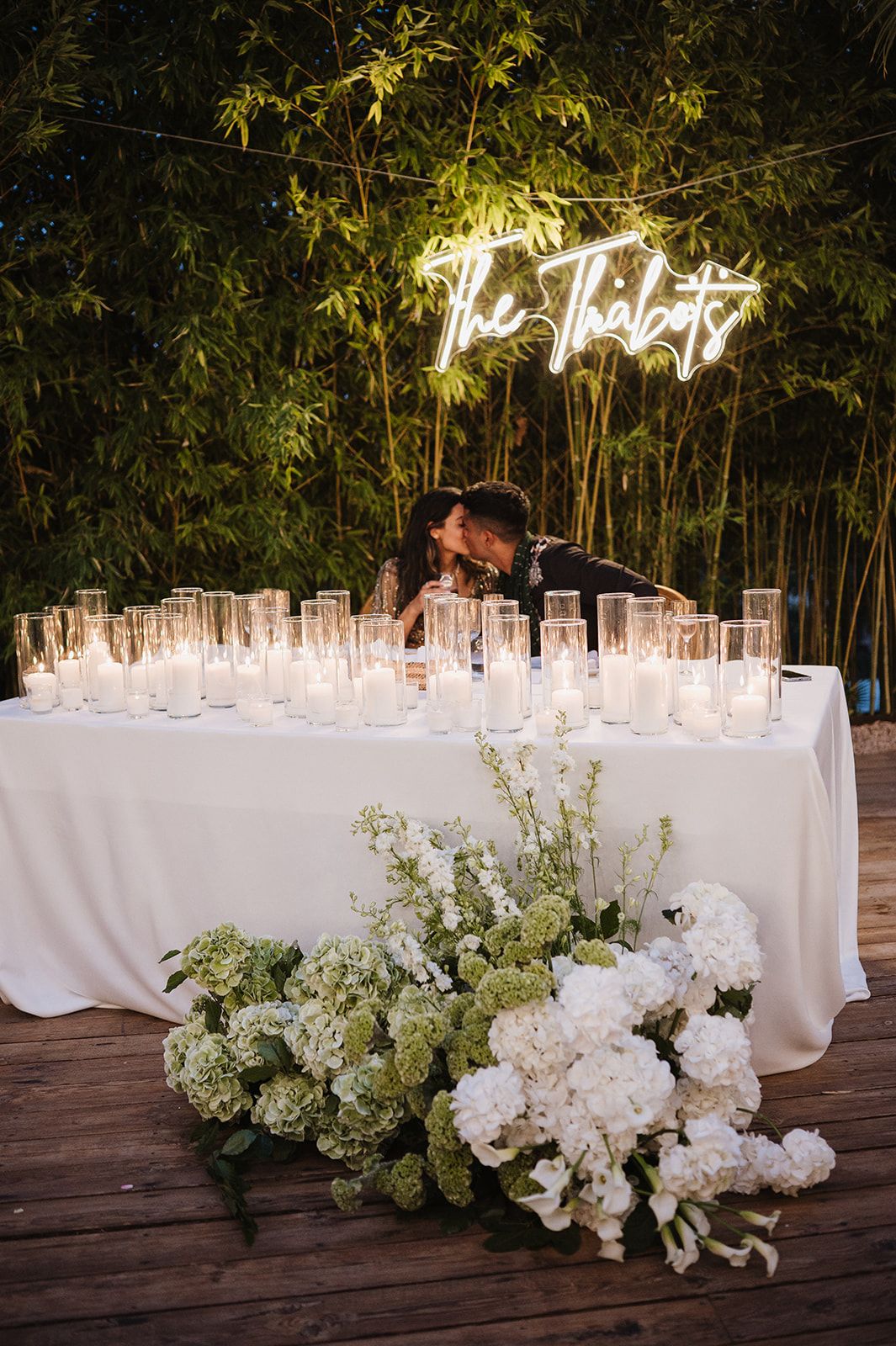 Couple kissing behind sweetheart table with candles, white florals, and glowing neon sign.