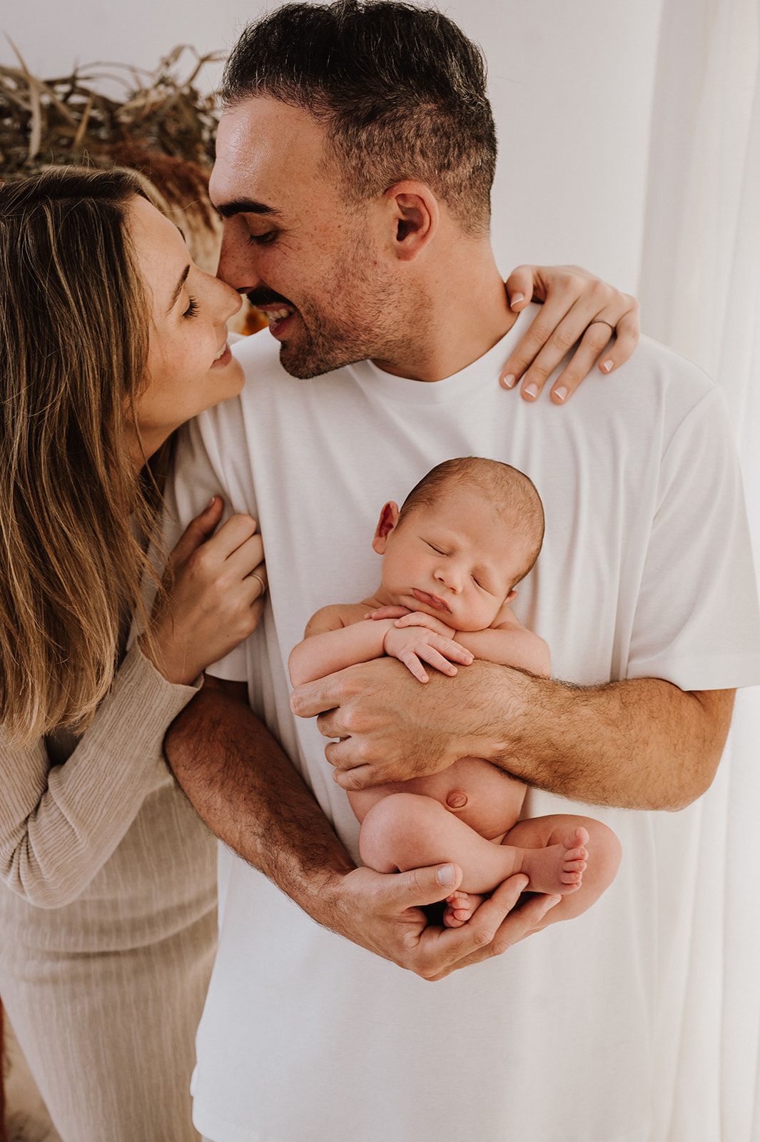 A couple smiles and kisses while the man gently cradles their sleeping newborn baby in his arms.