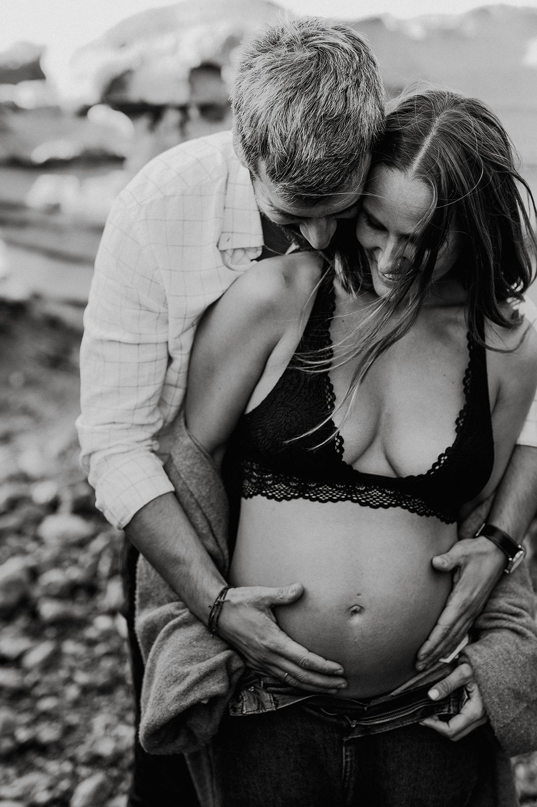 Black and white photo of a couple embracing outdoors, man kissing pregnant womans head while cradling her baby bump.