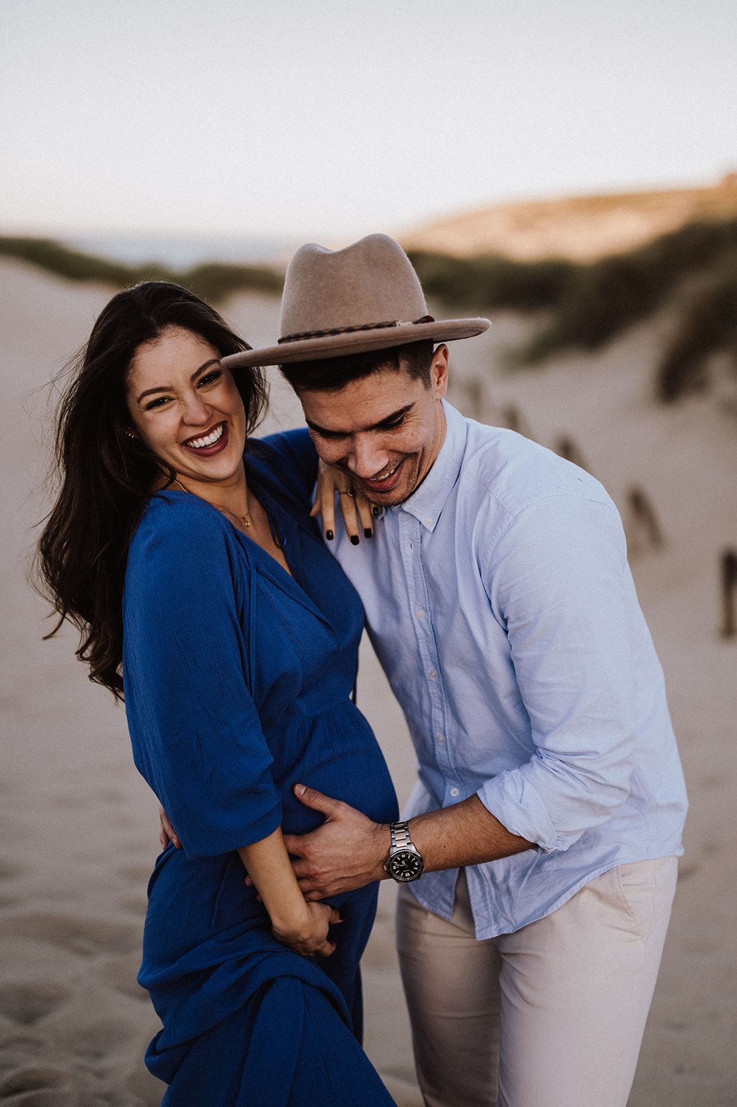 A laughing couple embraces on sandy dunes, woman in blue dress, man in tan hat and white shirt.