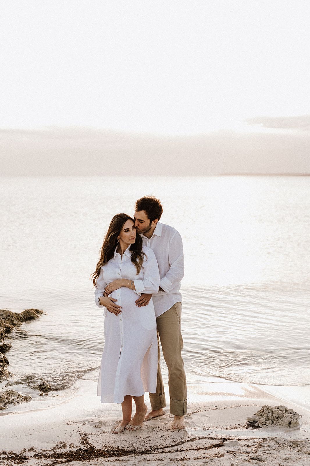 Couple in white outfits sharing a tender moment on a sandy beach, woman cradling baby bump during maternity photoshoot.