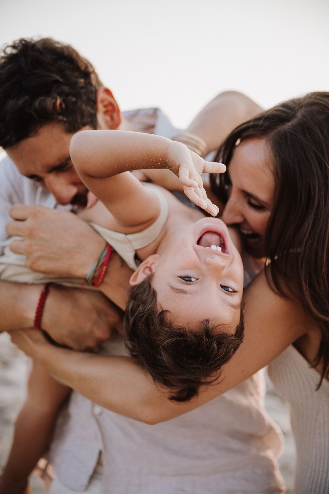 Two parents playfully hold their laughing baby upside down during a fun family photo session outdoors.