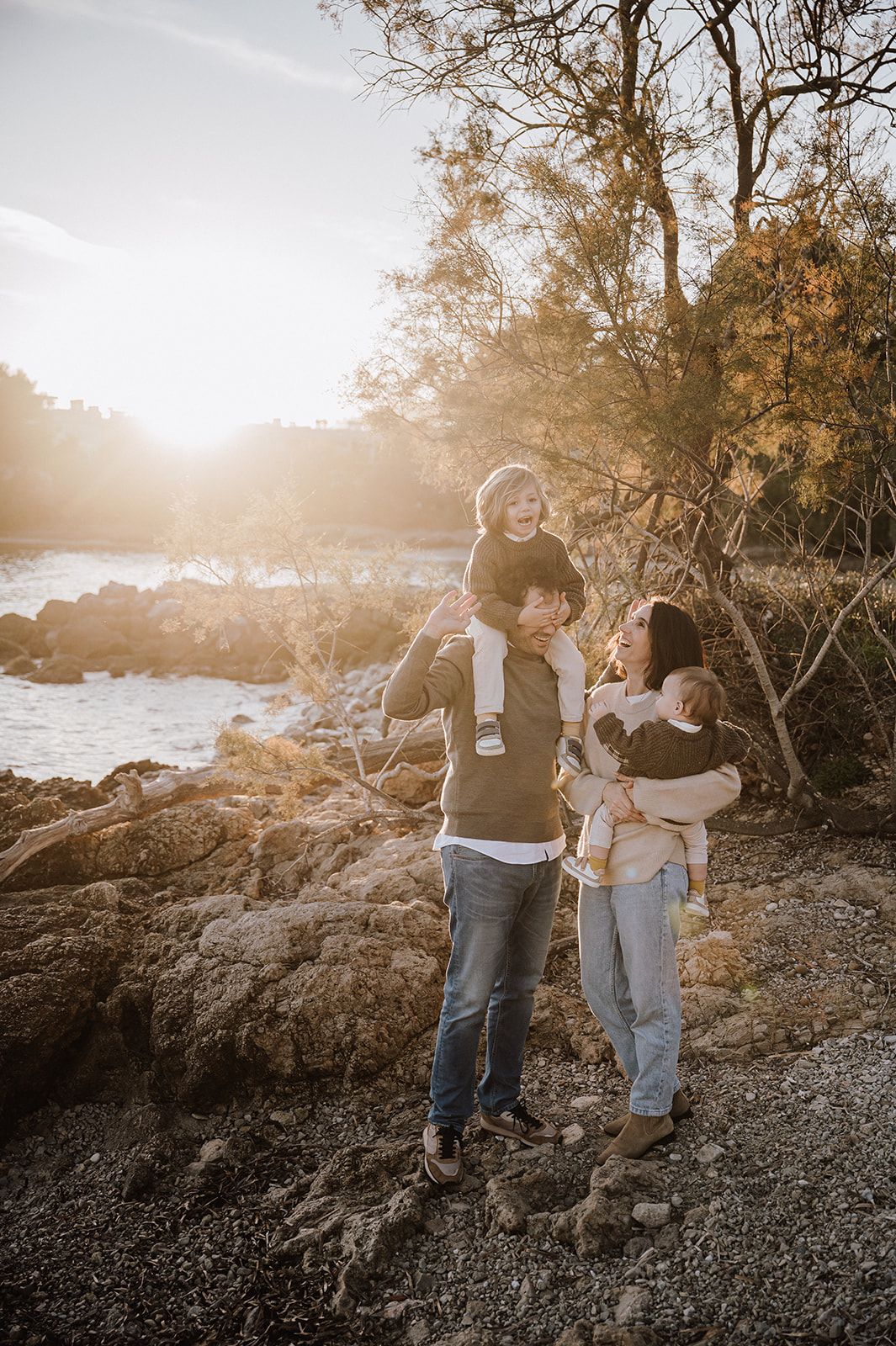 Family of four enjoying golden hour by a rocky shoreline, parents smiling as child sits on dad's shoulders.