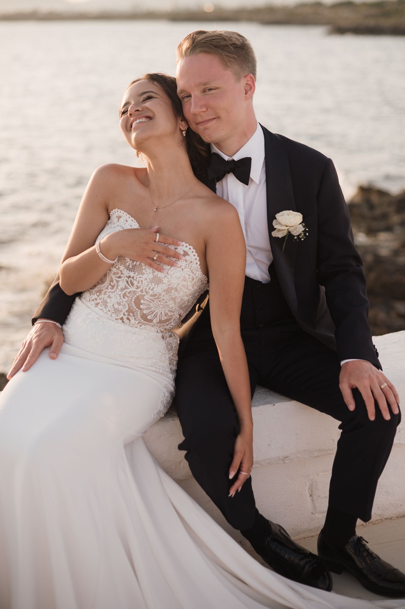 Bride and groom share romantic moment seated by the ocean at golden hour.