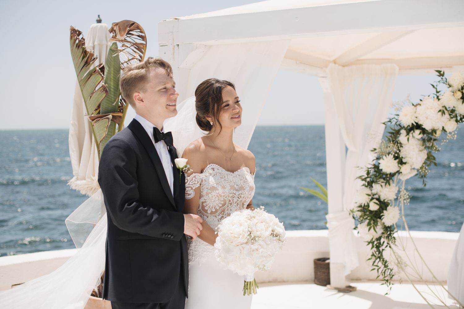 Smiling bride and groom stand at oceanfront wedding altar adorned with white floral arrangements.