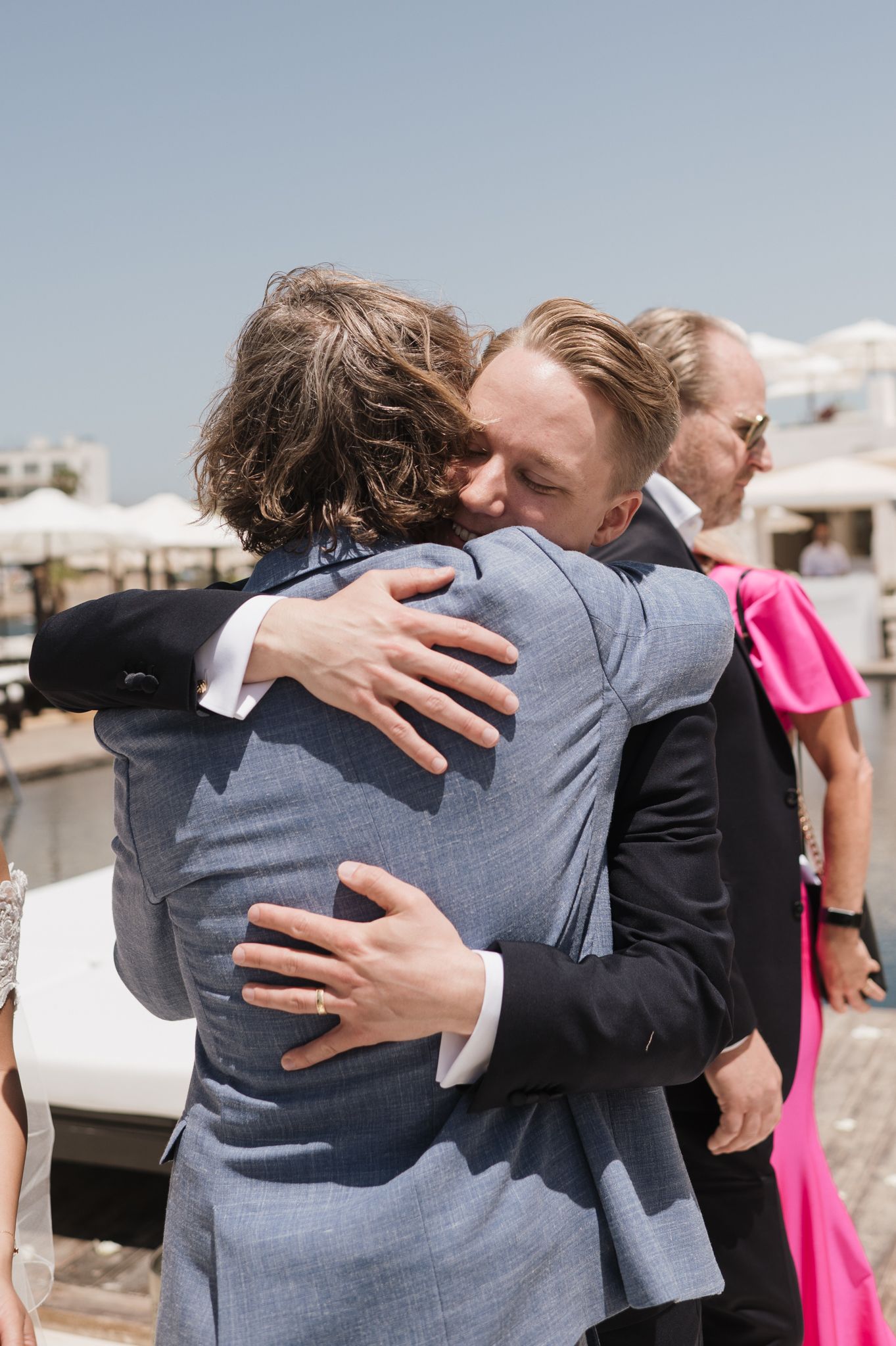 Groom in grey suit shares warm embrace with guest at outdoor waterfront wedding celebration.