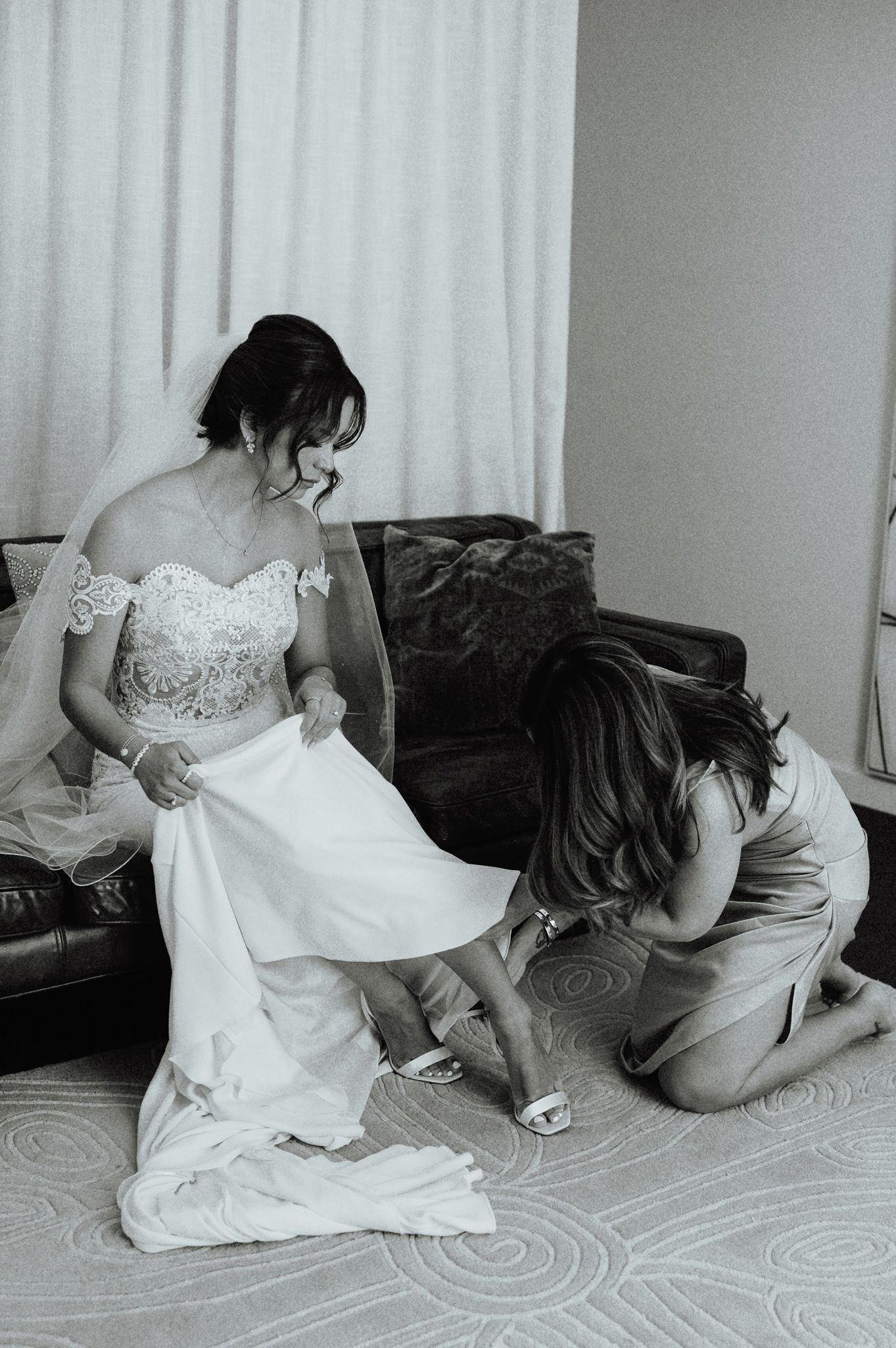 Bride in lace off-shoulder gown gets help with shoes from bridesmaid in black and white wedding photo.