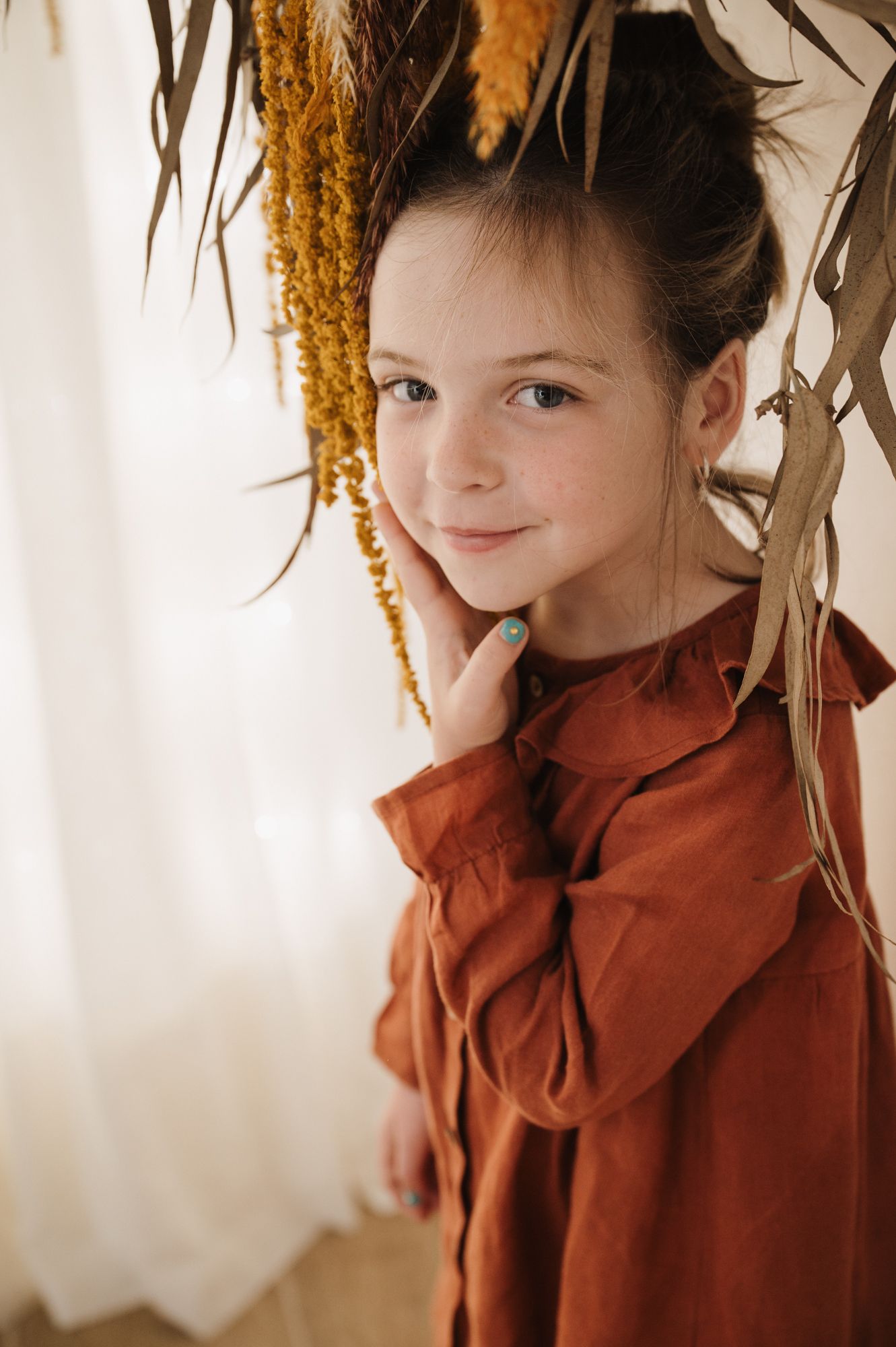Young girl in rust dress poses beside dried autumn botanicals, smiling softly at camera.