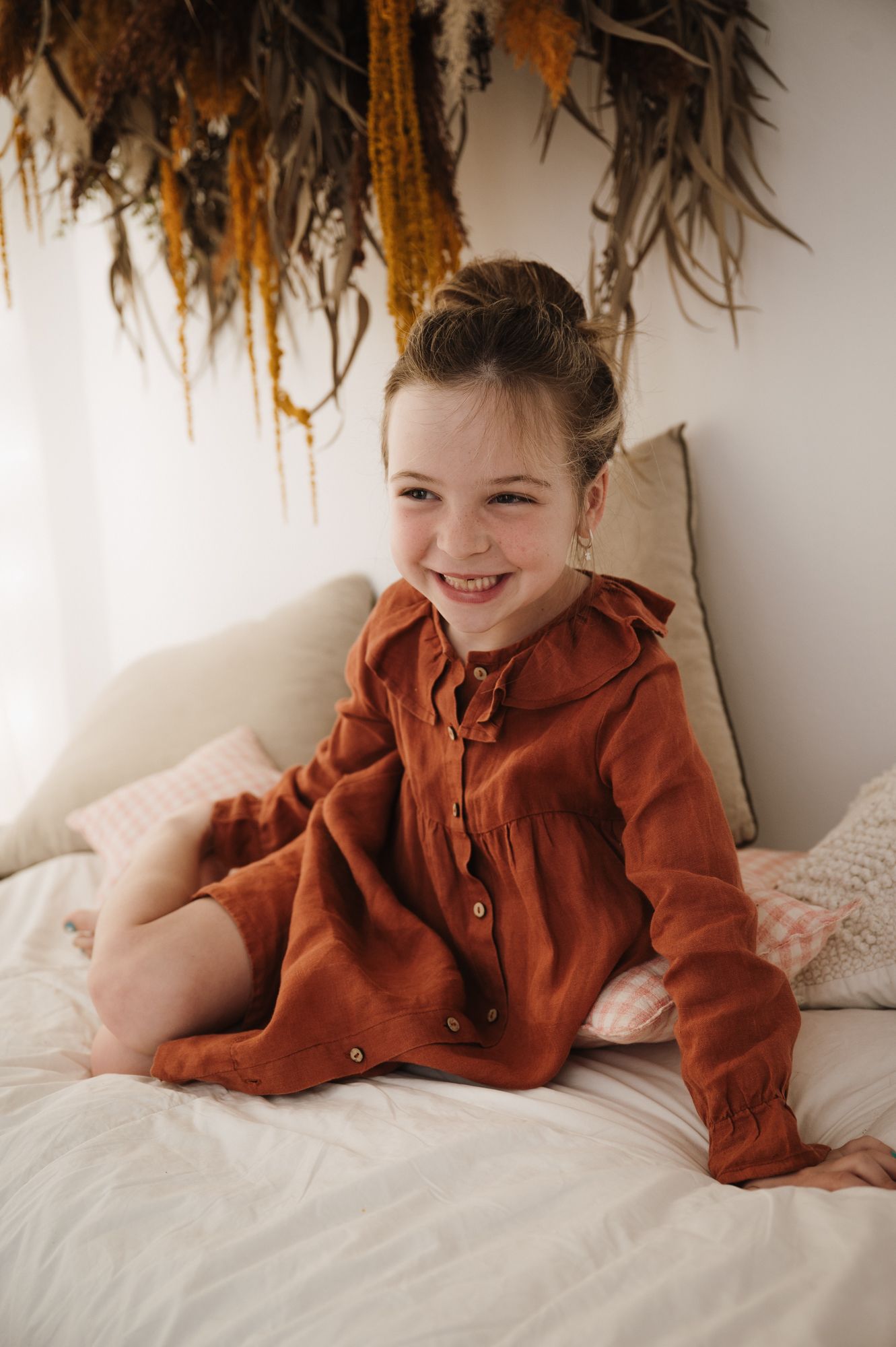 Smiling young girl in rust-colored linen dress sitting on white bed with dried floral wall decor.