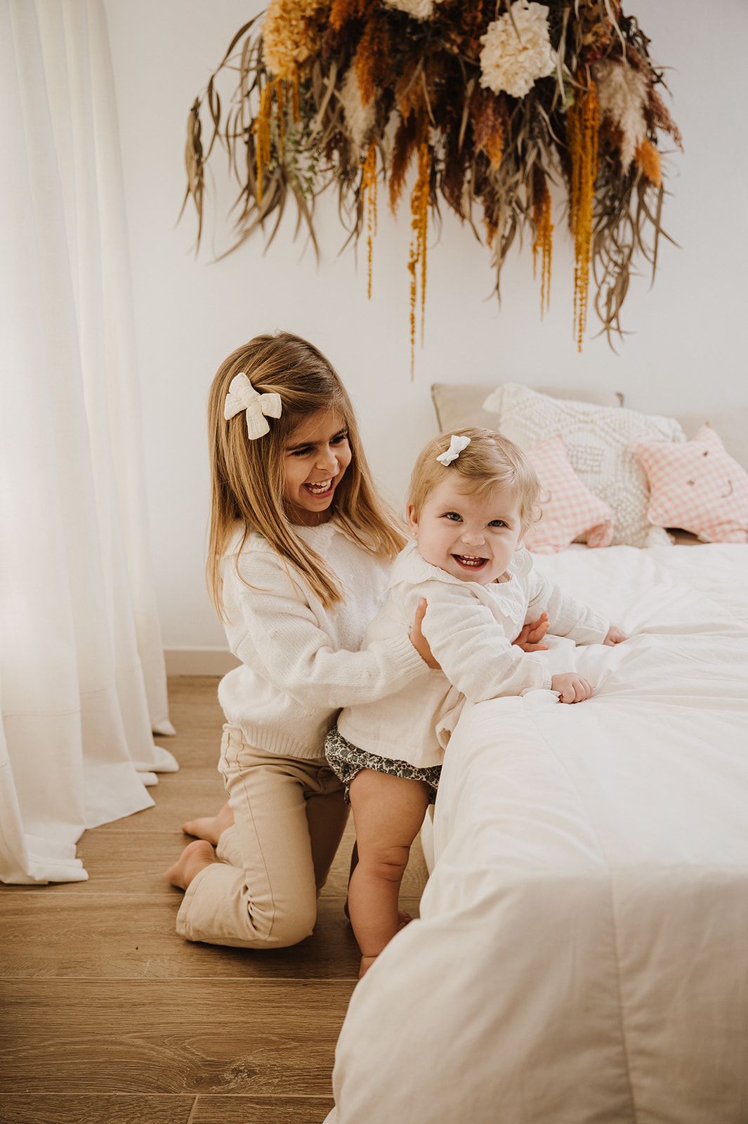A young girl smiles while helping a happy toddler stand beside a bed in a bright, cozy bedroom with floral decor.