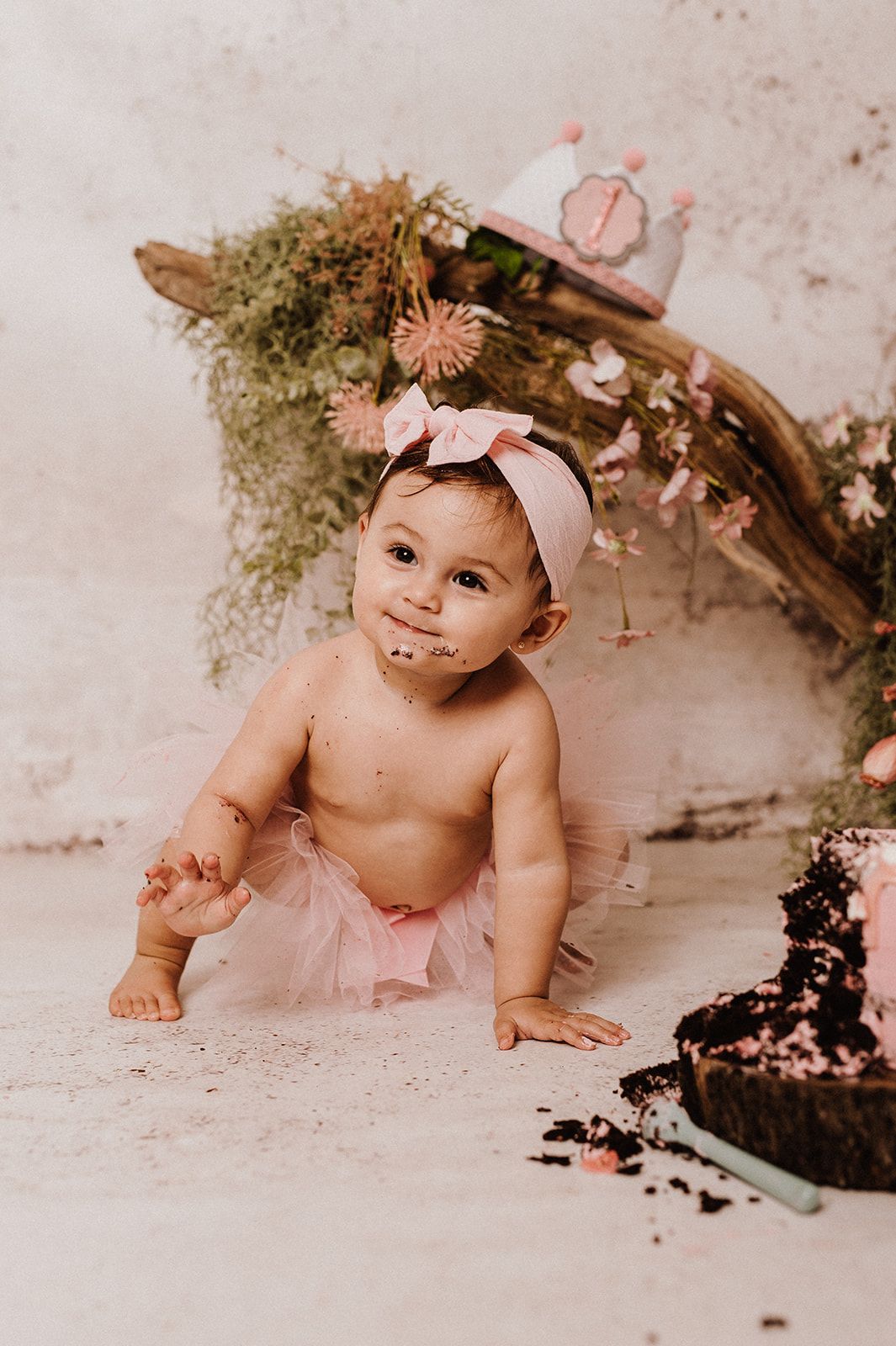 Baby girl in pink tutu and bow headband smashes first birthday cake during a floral cake smash photoshoot.