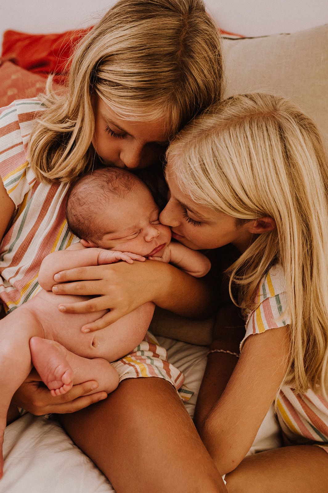 Two blonde sisters lovingly kiss and cuddle their newborn baby sibling while sitting together on a cozy bed.