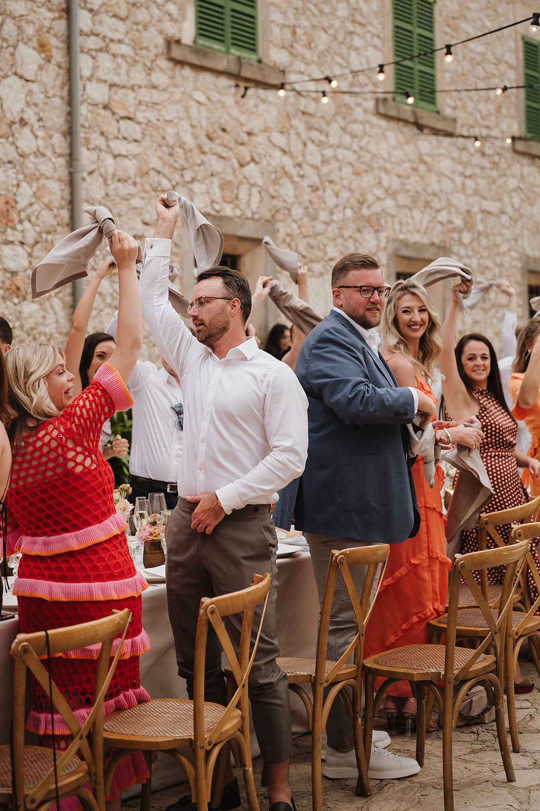 Joyful wedding guests waving napkins at outdoor stone venue reception with string lights.