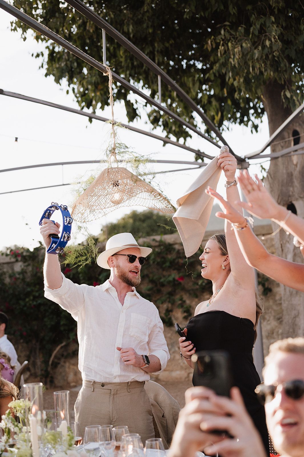 Energetic wedding guest celebrates at outdoor reception, playing tambourine and dancing under string lights.