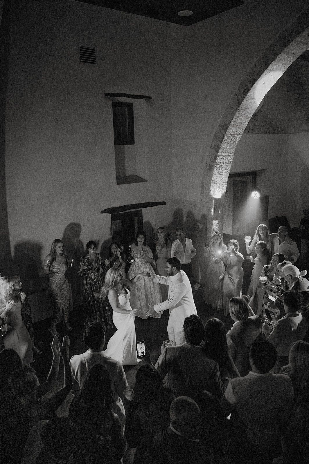 Black and white photo of bride and groom sharing first dance surrounded by wedding guests.