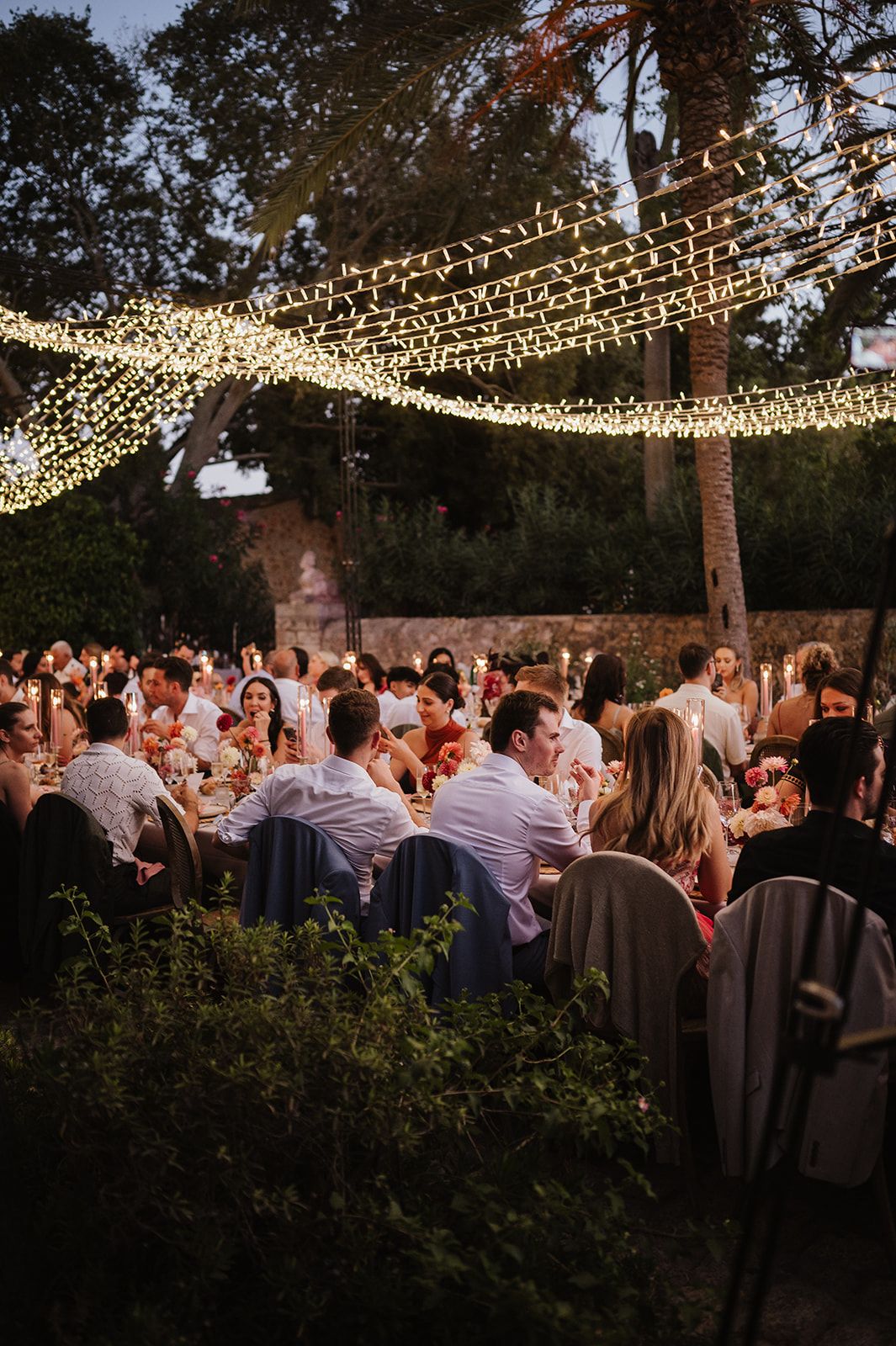 Wedding guests dine under a canopy of fairy lights at an enchanting outdoor evening reception.