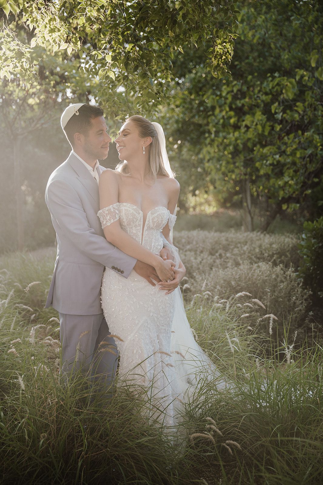 Bride and groom share tender moment in golden sunlit garden surrounded by tall grass.
