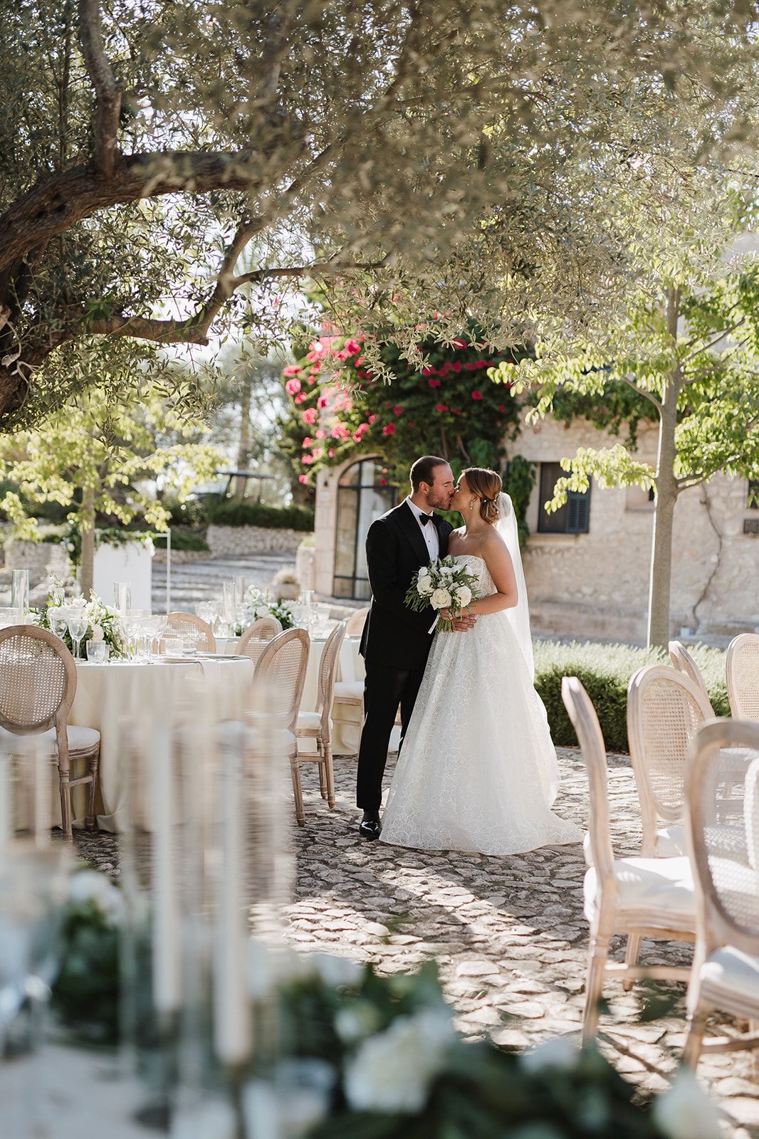 Newlyweds share a romantic kiss under an olive tree beside elegantly set outdoor reception tables.