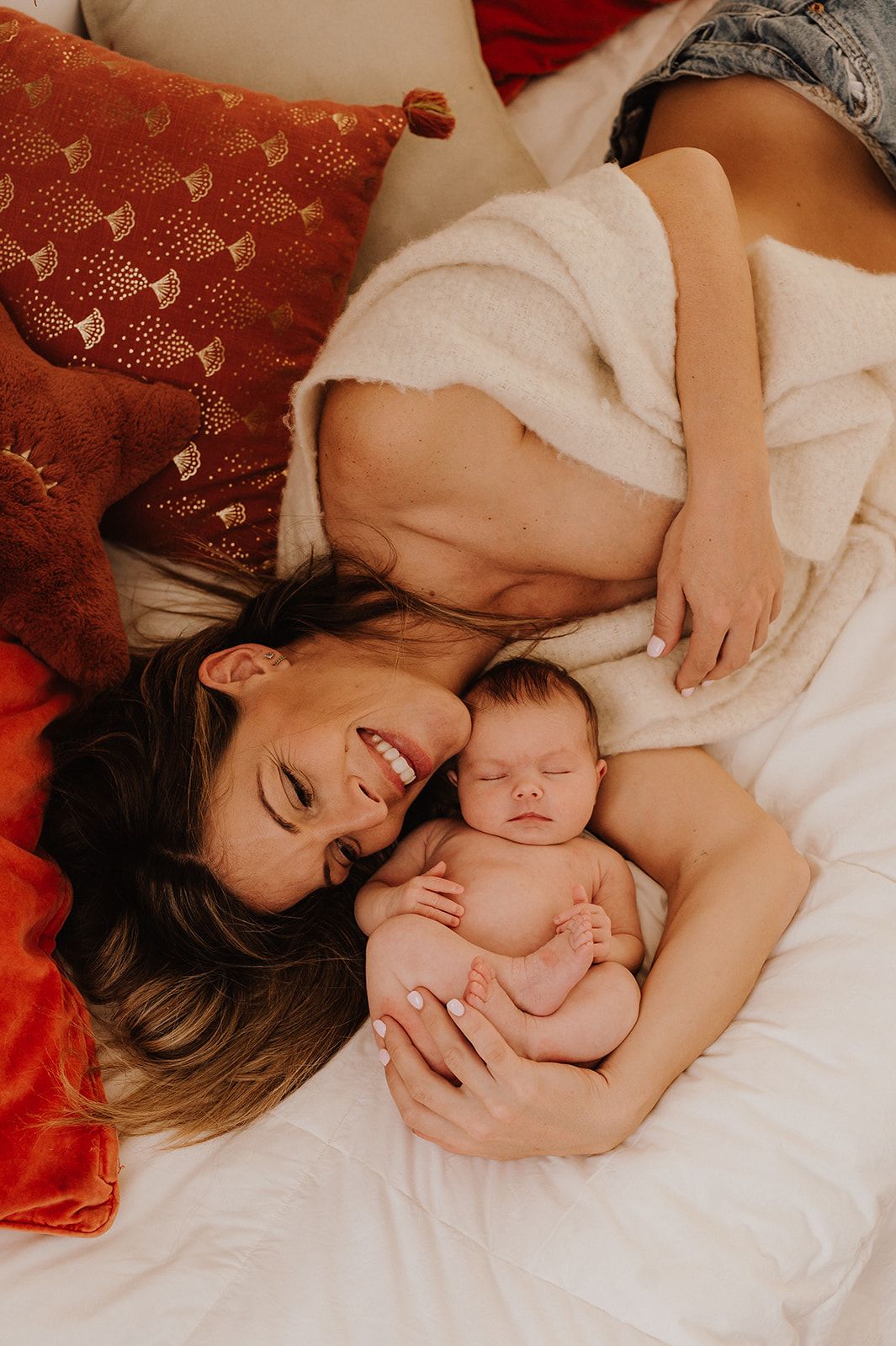 Smiling mother lying on white bed with sleeping newborn baby surrounded by red and orange decorative pillows.