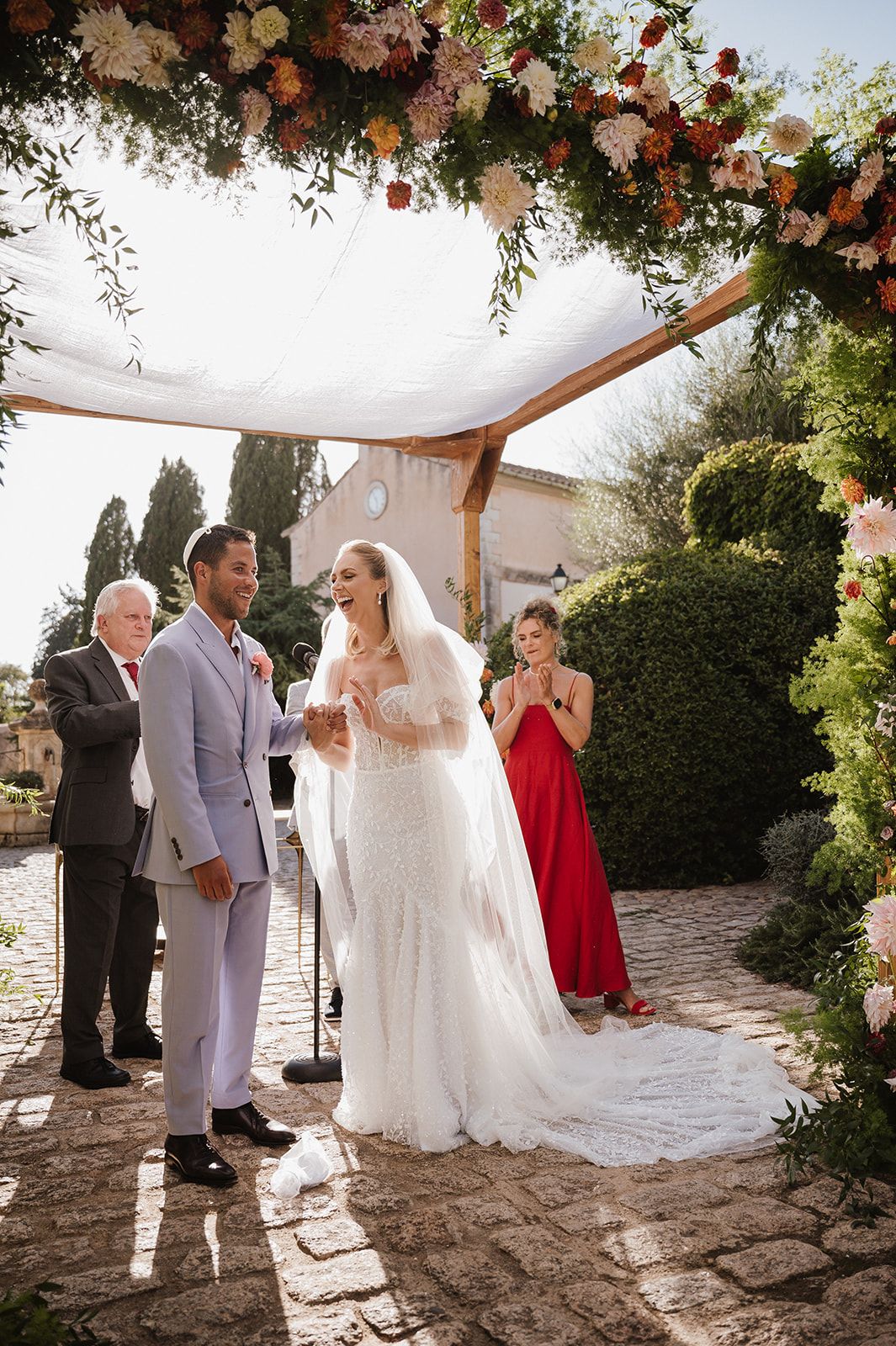 Bride and groom stand under floral chuppah during outdoor Jewish wedding ceremony in sunny garden.